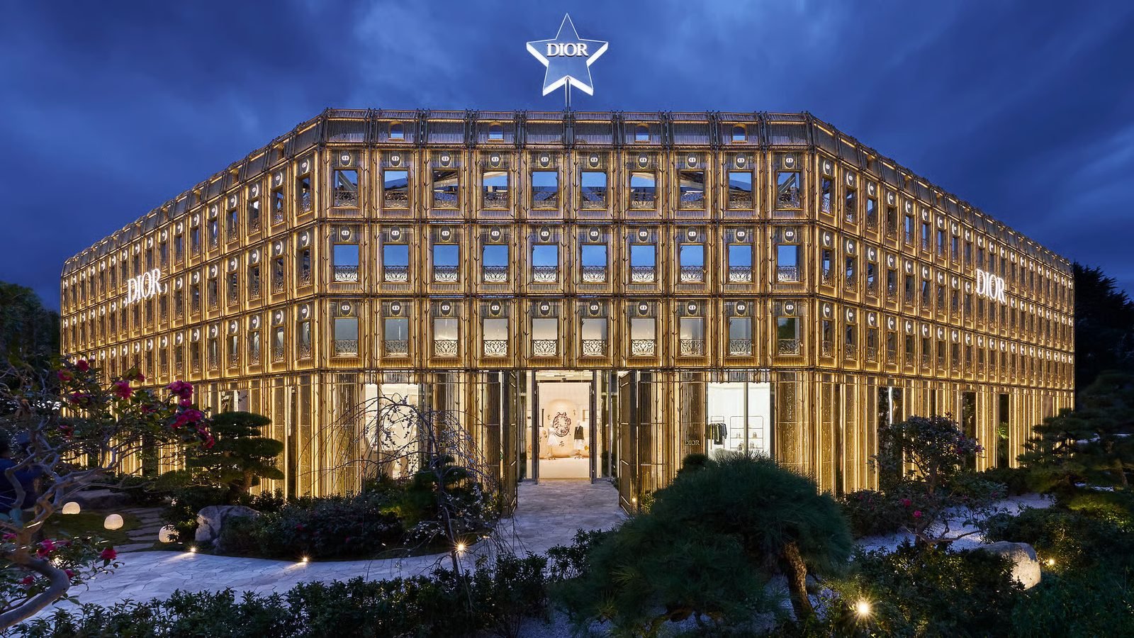 Fachada iluminada del Dior Bamboo Pavilion en Tokio al anochecer, con columnas doradas que imitan cañas de bambú y la estrella Dior en lo alto.